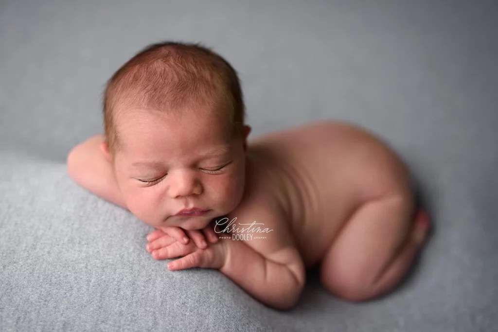A newborn baby is lying on their stomach on a soft, grey blanket. The baby is nude and sleeping peacefully with their hands folded under their chin and head slightly turned to the side. The background is out of focus, keeping the attention on the child. Image by Denver Newborn Photographer Christina Dooley Photography.