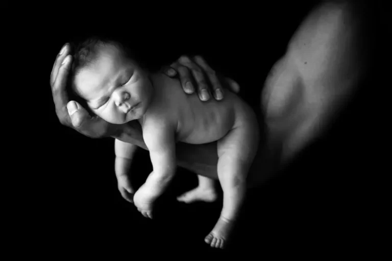 A black-and-white image of a newborn baby cradled gently in an adult's hands. The baby is asleep, with a peaceful expression. The surrounding background is dark, making the baby the focal point of the image—a timeless moment captured by Christina Dooley Photography, a Colorado Newborn Photographer. Image by Denver Newborn Photographer Christina Dooley Photography.