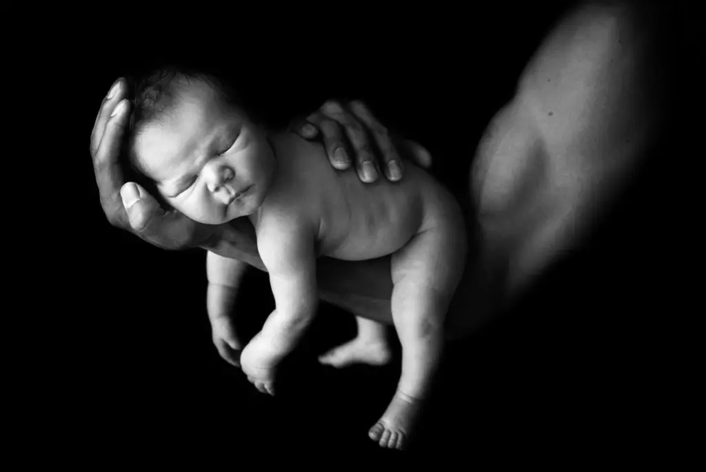 A black-and-white image of a newborn baby cradled gently in an adult's hands. The baby is asleep, with a peaceful expression. The surrounding background is dark, making the baby the focal point of the image—a timeless moment captured by Christina Dooley Photography, a Colorado Newborn Photographer. Image by Denver Newborn Photographer Christina Dooley Photography.