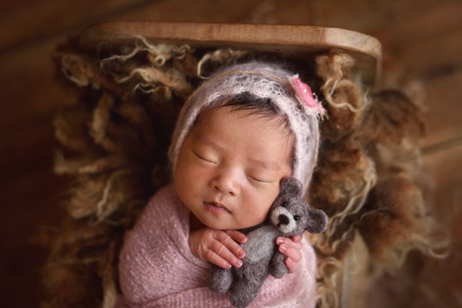 A newborn baby, swaddled in a pink blanket and wearing a knitted bonnet with a small flower, sleeps peacefully while holding a small, gray, stuffed teddy bear. This is the cutest baby you'll ever see in newborn photography, lying serenely on a textured, brown furry blanket. Image by Denver Newborn Photographer Christina Dooley Photography.