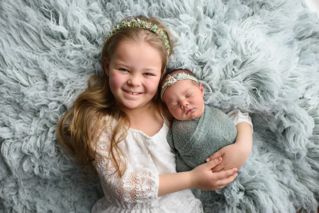 A young girl with long blonde hair and a flower headband smiles while lying on a fluffy, light grey blanket, holding a sleeping baby swaddled in a green wrap who also has a matching headband. Both children are dressed in white and appear content. Image by Denver Newborn Photographer Christina Dooley Photography.