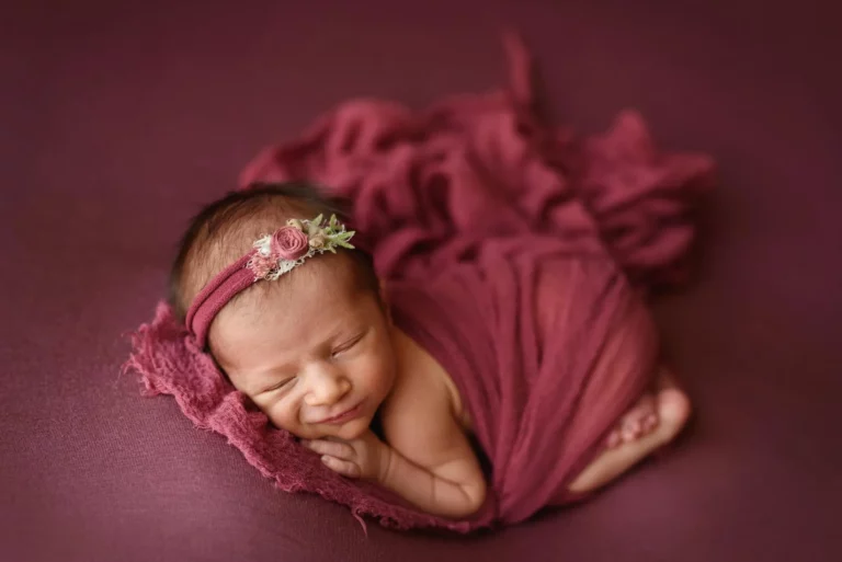 A newborn baby is peacefully sleeping, wrapped in a soft, maroon blanket. The baby wears a delicate headband adorned with small flowers. The background is also maroon, creating a warm and cozy atmosphere. The baby seems to be smiling gently in sleep. Image by Denver Newborn Photographer Christina Dooley Photography.