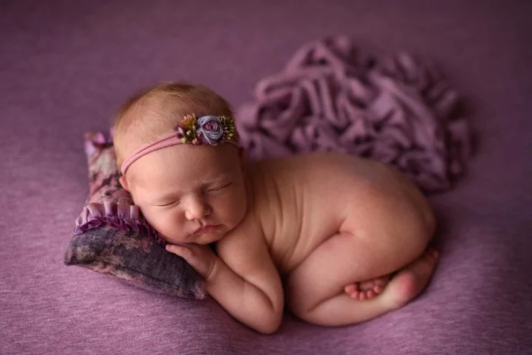 An infant peacefully sleeping on their side on a purple blanket, wearing a purple headband with a decorative flower. The baby is using a small purple pillow for support, and there is ruffled purple fabric in the background. Captured beautifully by Christina Dooley Photography, Colorado Newborn Photographer. Image by Denver Newborn Photographer Christina Dooley Photography.