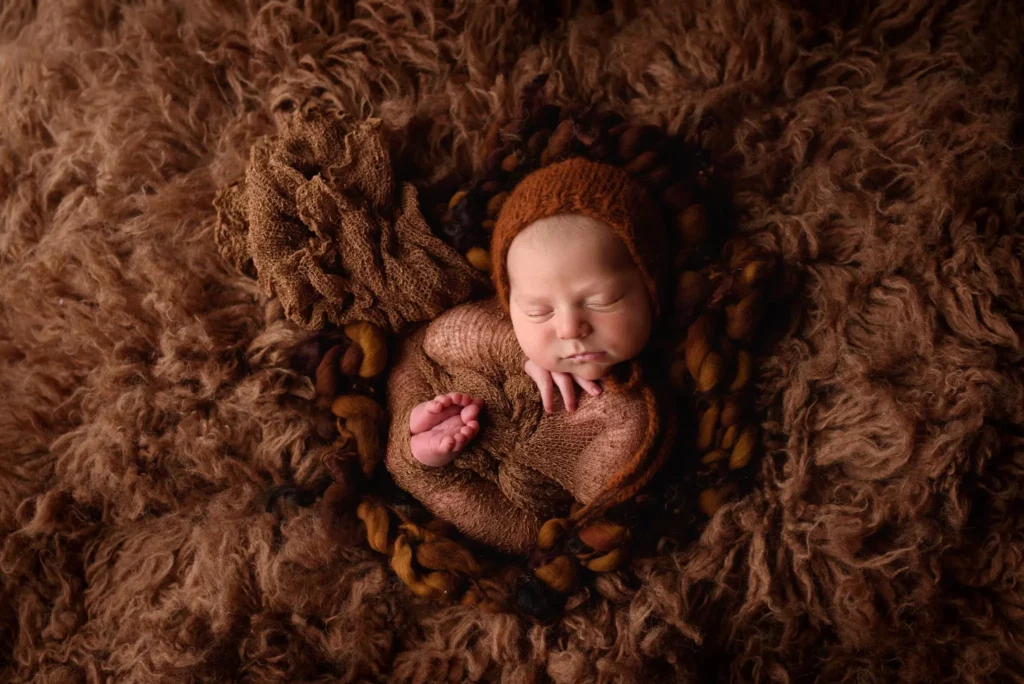 A sleeping newborn baby is wrapped in a brown knit blanket and wearing a matching brown bonnet. The baby is nestled in a fluffy, textured brown backdrop, surrounded by soft, earthy-colored layers, creating a cozy and warm atmosphere. Image by Denver Newborn Photographer Christina Dooley Photography.