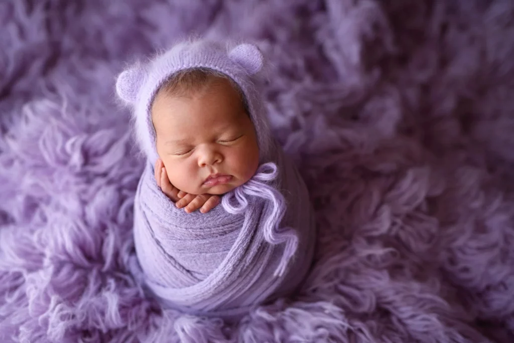 A sleeping baby is swaddled in a soft purple wrap and wears a matching hat with bear ears, surrounded by a fluffy, purple blanket. The baby has a serene expression and is resting with one hand under their chin. Image by Denver Newborn Photographer Christina Dooley Photography.