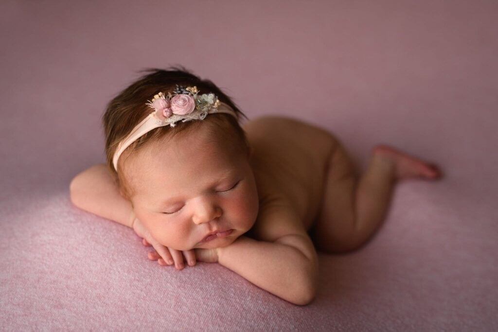 A newborn baby sleeps peacefully on a soft, pink blanket, embodying the pure joy of postpartum life. Resting on their stomach with hands folded under their chin, the baby wears a delicate headband adorned with small flowers, adding a touch of sweetness to the serene scene. Image by Denver Newborn Photographer Christina Dooley Photography.