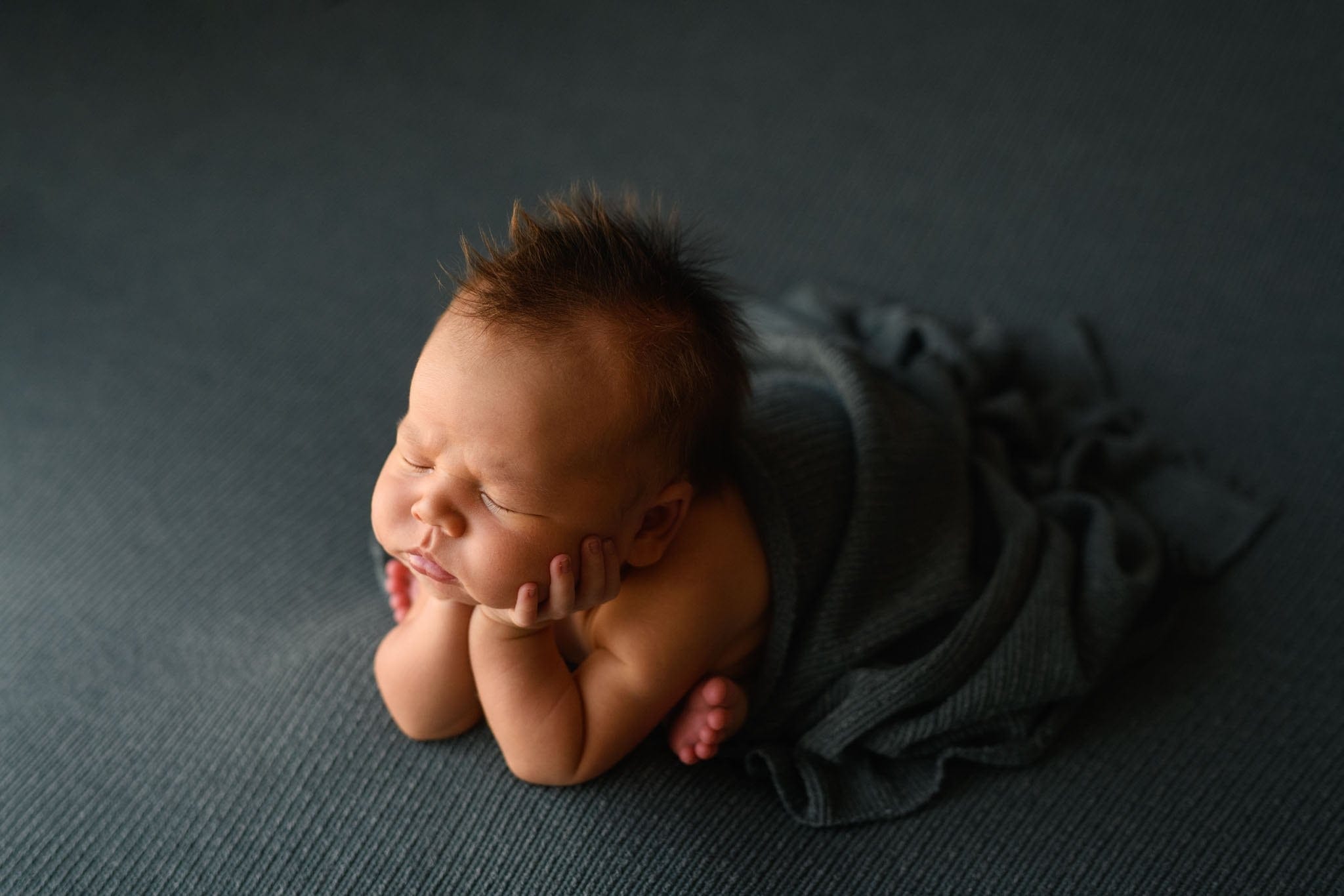 A newborn baby is asleep with their head resting on their hands, lying on a dark, textured surface. Swaddled in a dark gray blanket from Bumps and Bundles, their hair styled in a slight mohawk. The scene is softly lit, creating a peaceful atmosphere that highlights the perfect photo outfits for such precious moments. Image by Denver Newborn Photographer Christina Dooley Photography.