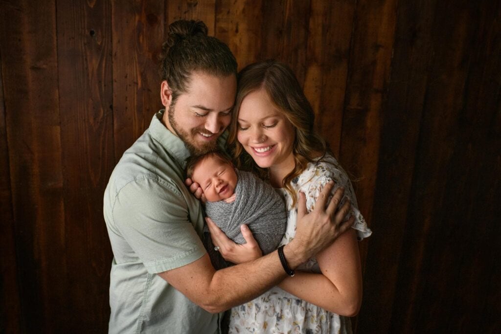 A couple lovingly embraces their sleeping newborn. The father, with a beard and tied-back hair, holds the baby swaddled in a gray blanket, while the smiling mother with wavy hair gently supports the child's head. They all stand in front of a dark wooden backdrop, reflecting their beautiful birth experience. Image by Denver Newborn Photographer Christina Dooley Photography.