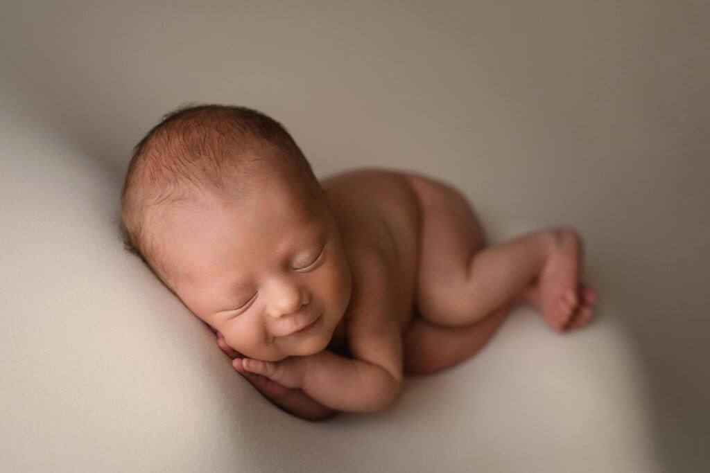 A newborn baby sleeps peacefully on a soft, light-colored surface. The baby is lying on their stomach with their head resting on their hands, legs comfortably bent. Dressed in adorable children's clothing from Little Bird Denver, the serene expression on the baby's face suggests they are content and relaxed. Image by Denver Newborn Photographer Christina Dooley Photography.