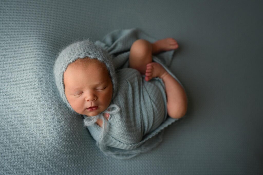 A newborn baby, swaddled in a light blue blanket and wearing a matching knit hat from *Once Upon a Child Denver*, sleeps peacefully on a textured blue background. The baby's arms and legs are tucked in, creating a serene and cozy scene reminiscent of *stylish kids clothes*. Image by Denver Newborn Photographer Christina Dooley Photography.