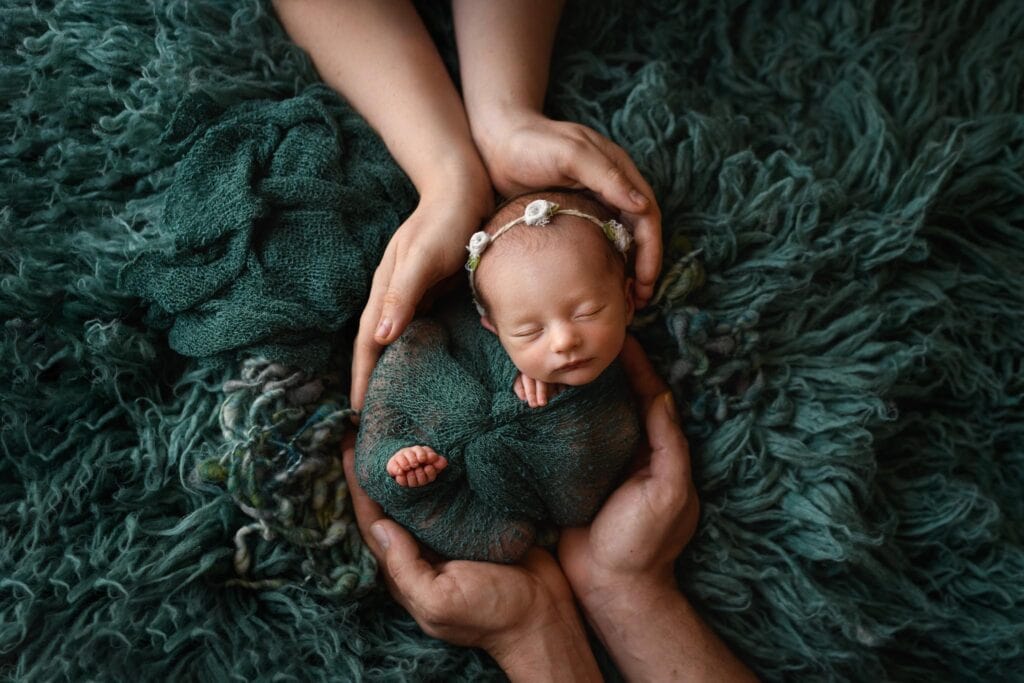 A newborn baby wrapped in green knit swaddle is gently cradled by the hands of mom and dad after using a Denver Midwife