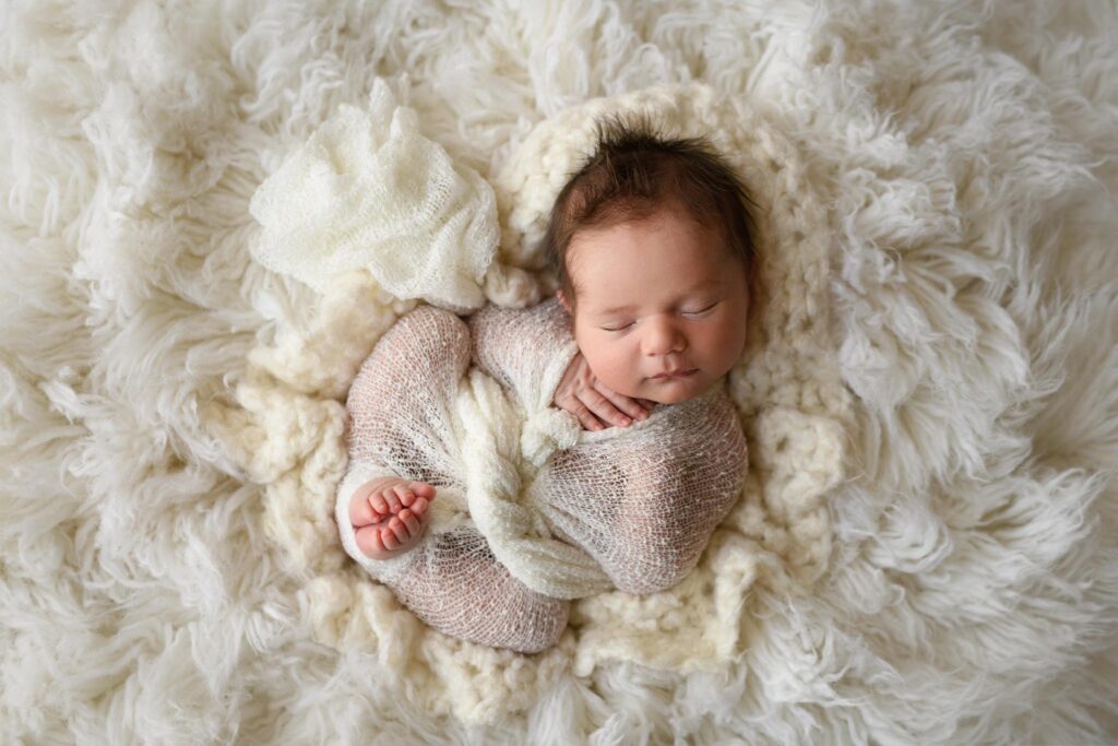 A newborn baby sleeps peacefully, wrapped in a soft white and beige knit blanket and nestled in a fluffy, cream-colored rug after visiting Broomtail Denver
